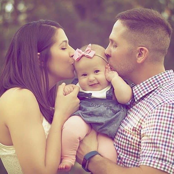 A man and woman kissing a baby's cheeks while holding her outdoors
